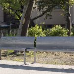 A modern metal bench with perforated backrest and armrests is placed on a paved path beside trees and a fenced area in a park.