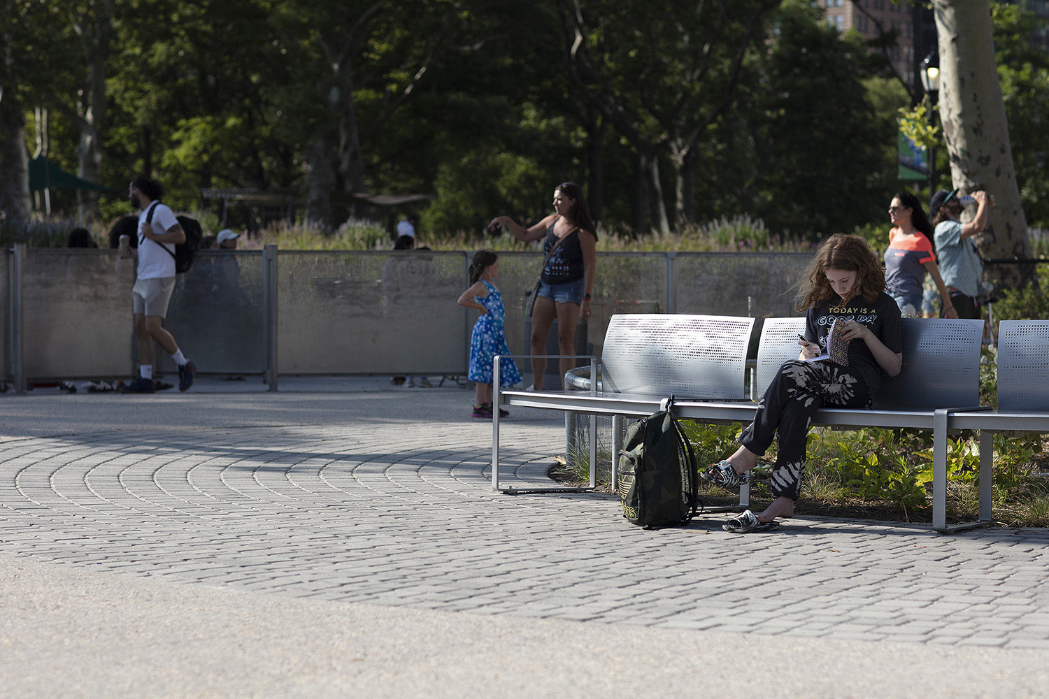 Une personne est assise sur un banc métallique et lit, un sac étant posé sur le sol à côté d'elle. D'autres personnes et des enfants se tiennent debout ou marchent à l'arrière-plan dans un parc.