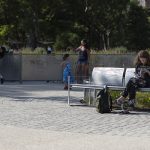 Une personne est assise sur un banc métallique et lit, un sac étant posé sur le sol à côté d'elle. D'autres personnes et des enfants se tiennent debout ou marchent à l'arrière-plan dans un parc.