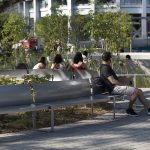 A man sits on a bench in an urban park while three people sit on another bench nearby, surrounded by trees and modern buildings.