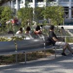 Several people sit on metal benches in a sunny, landscaped urban park area with greenery and modern buildings in the background.
