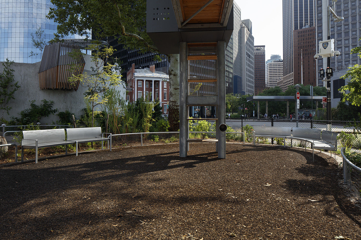 Small urban park area with benches, mulch ground, green plants, and modern metal structure, surrounded by city buildings and a sidewalk in the background.