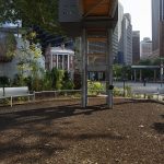 Small urban park area with benches, mulch ground, green plants, and modern metal structure, surrounded by city buildings and a sidewalk in the background.