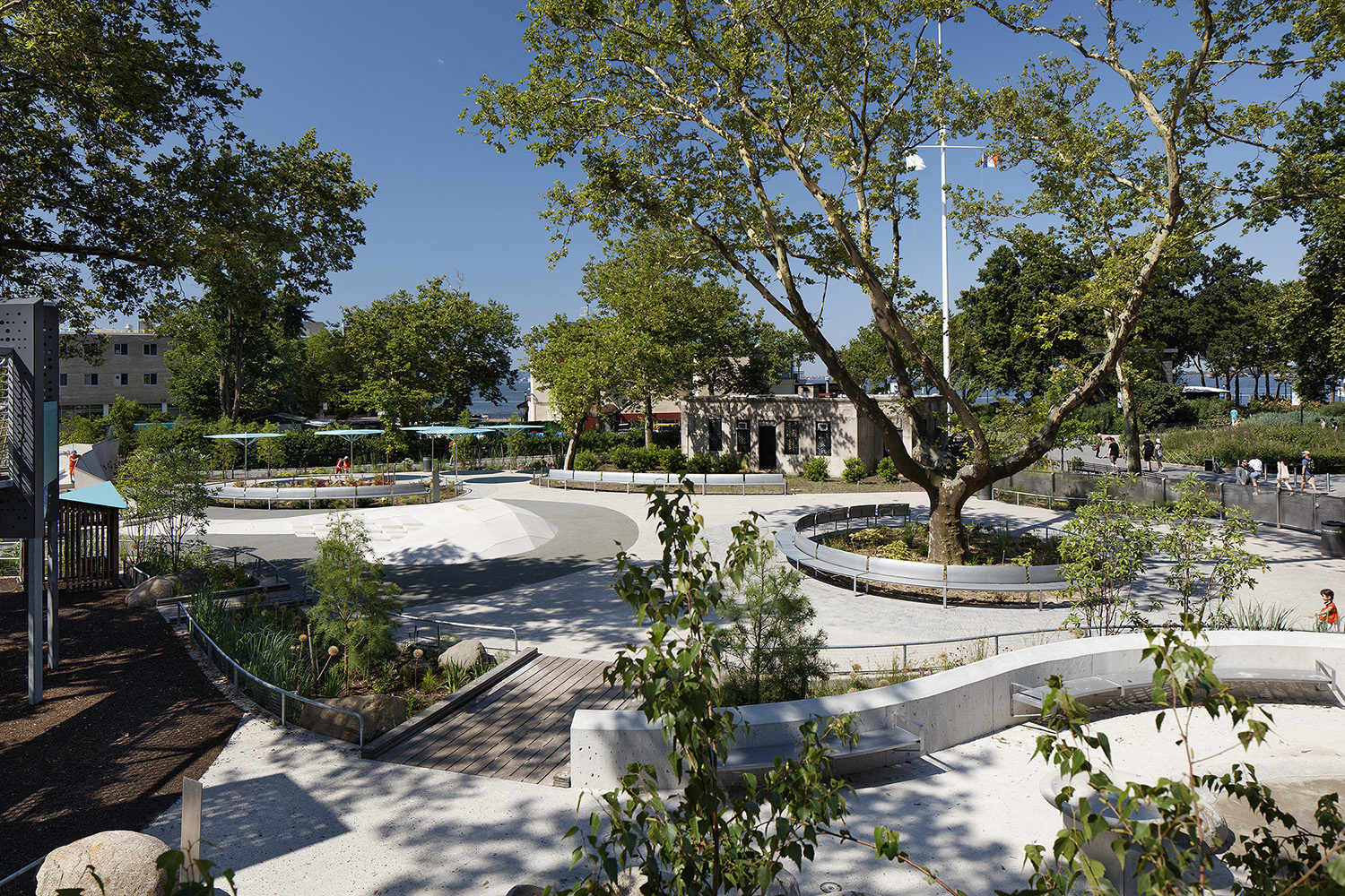 A landscaped urban park with curved walkways, seating areas, trees, and people walking on a sunny day.