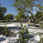 A landscaped urban park with curved walkways, seating areas, trees, and people walking on a sunny day.