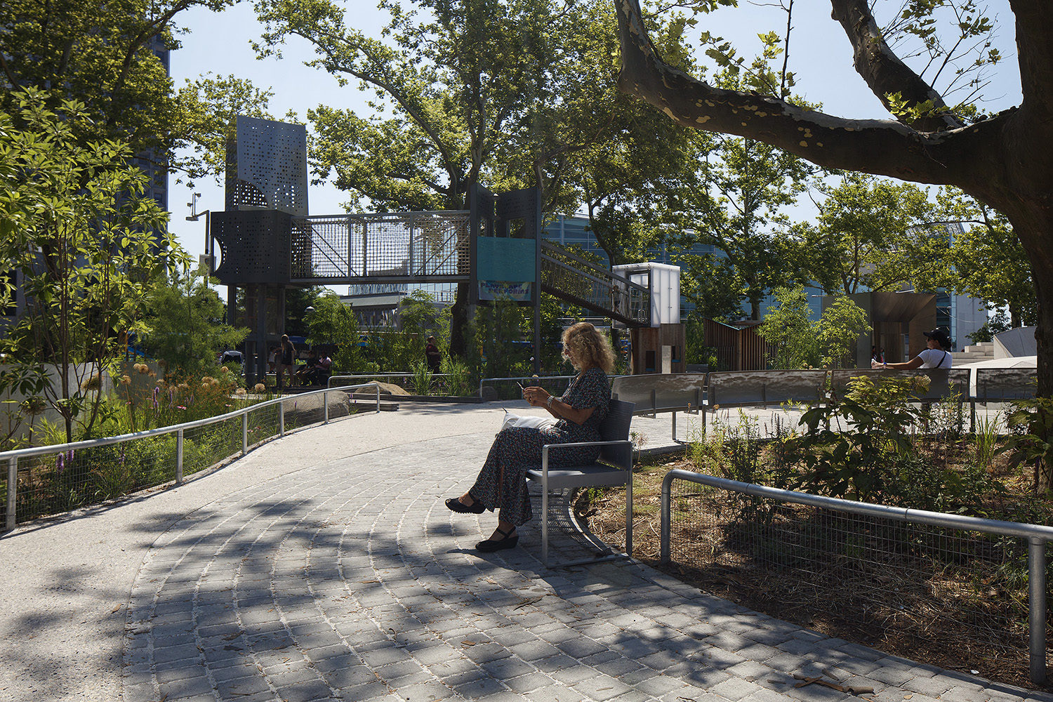 A person sits on a bench along a paved path in a sunlit park with trees, plants, and modern structures in the background.
