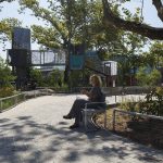 Une personne est assise sur un banc, lisant ou utilisant une tablette dans un parc paysager ombragé, avec des arbres, des chemins courbes et des structures modernes en arrière-plan.