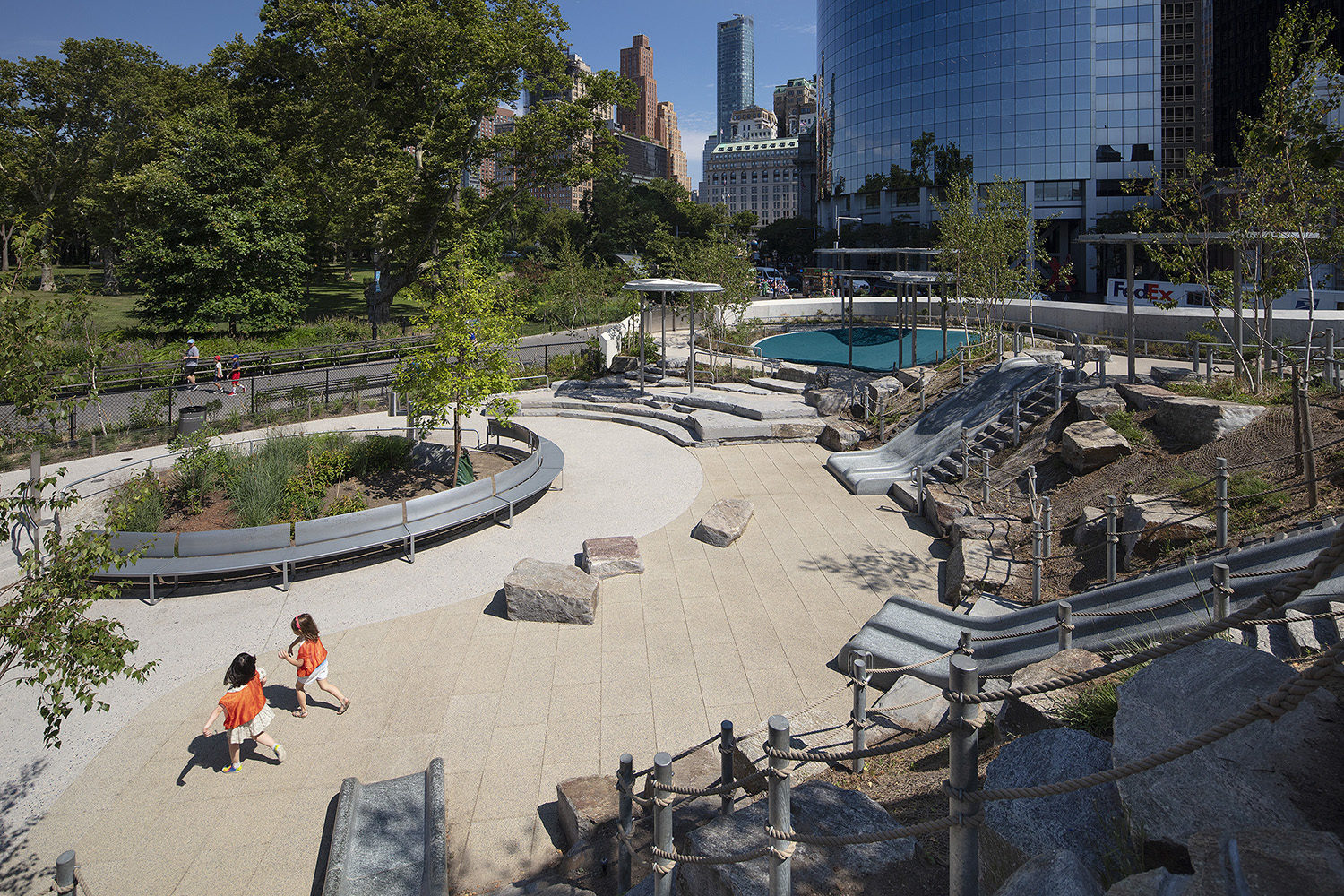 Two children run through an outdoor playground with slides, benches, trees, and nearby city buildings.