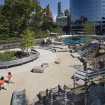 Two children run through an outdoor playground with slides, benches, trees, and nearby city buildings.