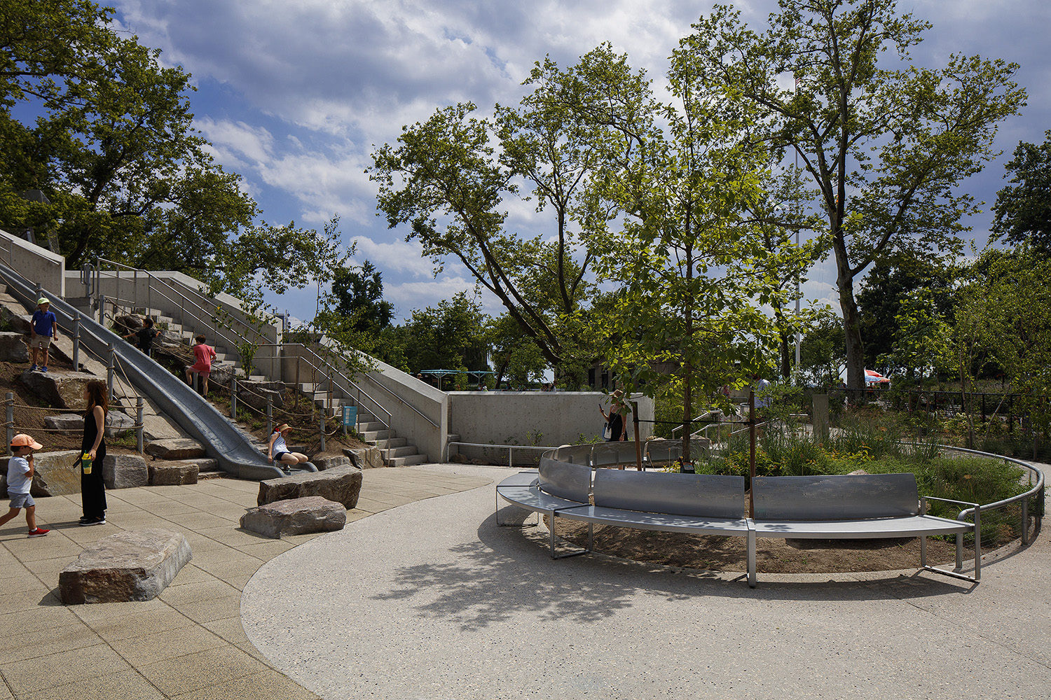 Children and adults are in a park featuring a playground with a slide, climbing rocks, and a circular bench around a tree, under a partly cloudy sky.
