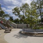 Aire de jeux en plein air avec un toboggan en métal, des marches en pierre, de gros rochers, des arbres, des bancs incurvés, et plusieurs personnes profitant de l'endroit par une journée partiellement nuageuse.