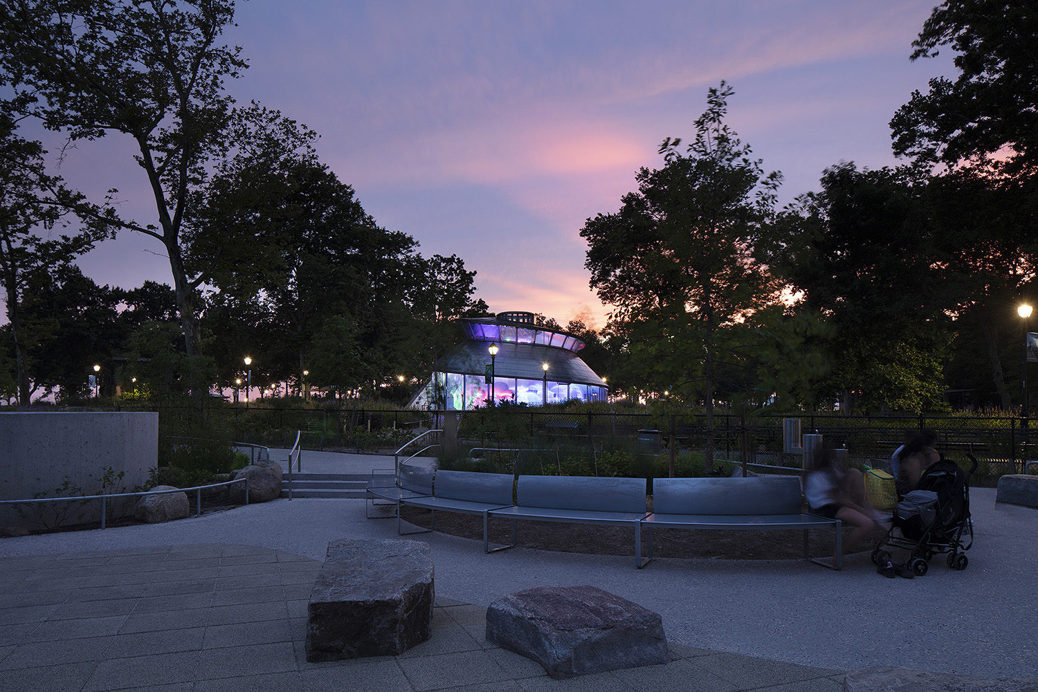 A modern glass pavilion is illuminated at dusk in a park, with trees and benches in the foreground and a person pushing a stroller.