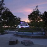 A modern glass pavilion is illuminated at dusk in a park, with trees and benches in the foreground and a person pushing a stroller.