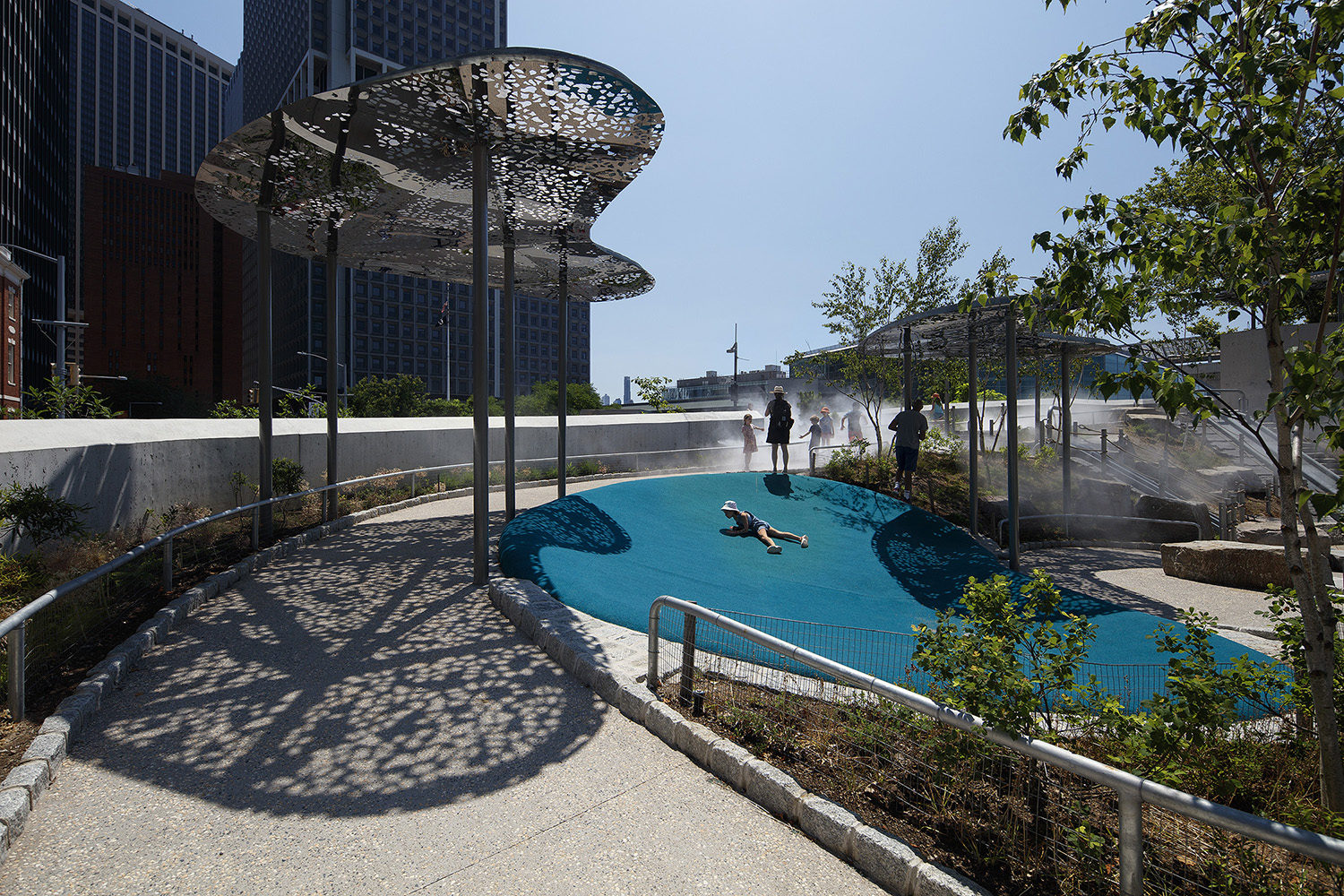 Lasercut cloud-shaped canopy structure casts a dappled shadow next to children playing at the Battery Playscape