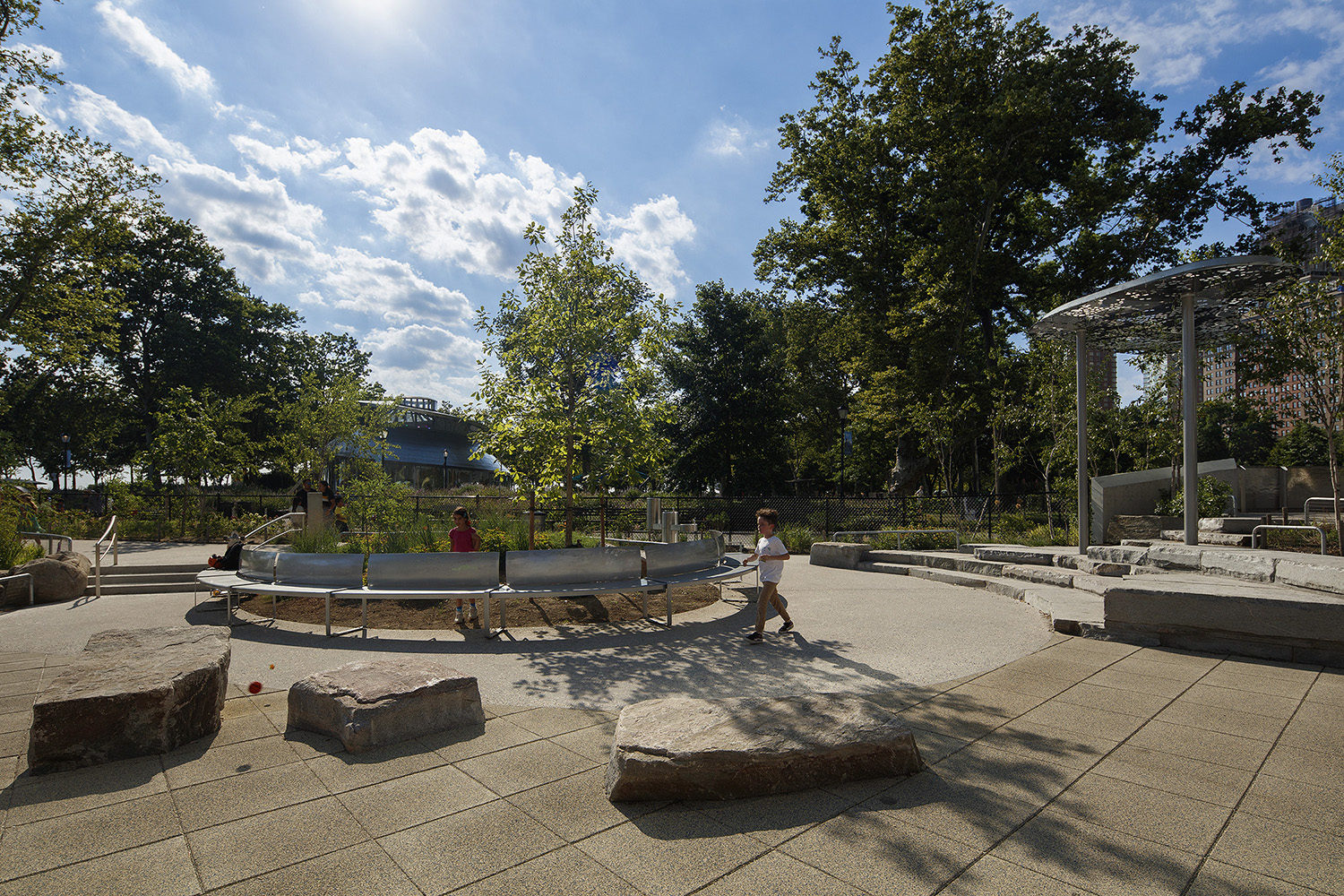 Battery-Playscape_shade-clouds_custom-curved-bench_001 Two children walk by metal benches at the Battery Playscape