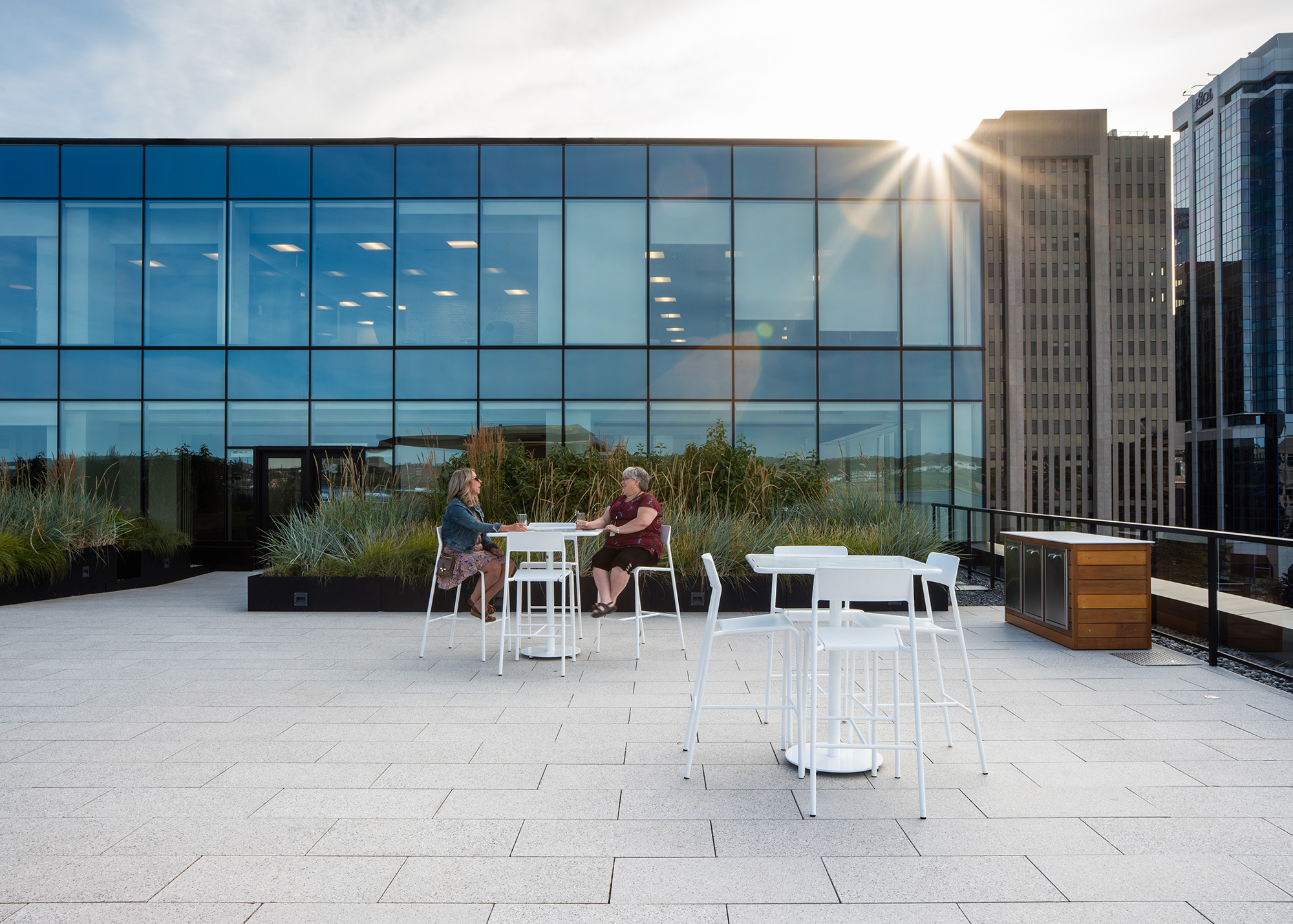 Foro-chairs-tables-stools_stewart-mckelvey_02 Two women chat while sitting on white Foro Bar height stools at a table in front of a large planter box