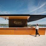 White Foro tables and chairs are on either side of the bar while a woman walks in the foreground of a rooftop terrace