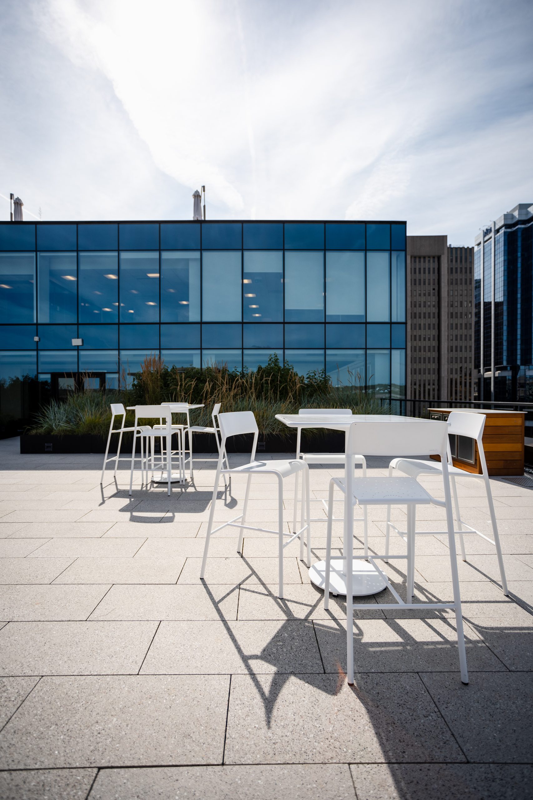 White outdoor tables and chairs on a rooftop patio with potted plants, in front of a modern glass building under a partly cloudy sky.