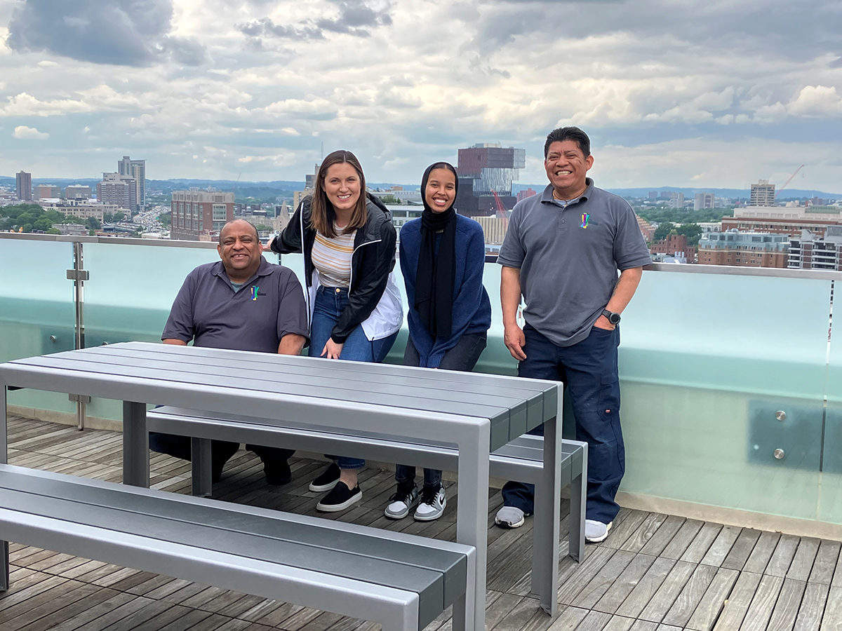viridian-1050-tables-benches-people Four maintenance workers at the Viridian Apartments smile pose on the roof deck with newly installed 1050 tables and benches. The Boston skyline is in the background.