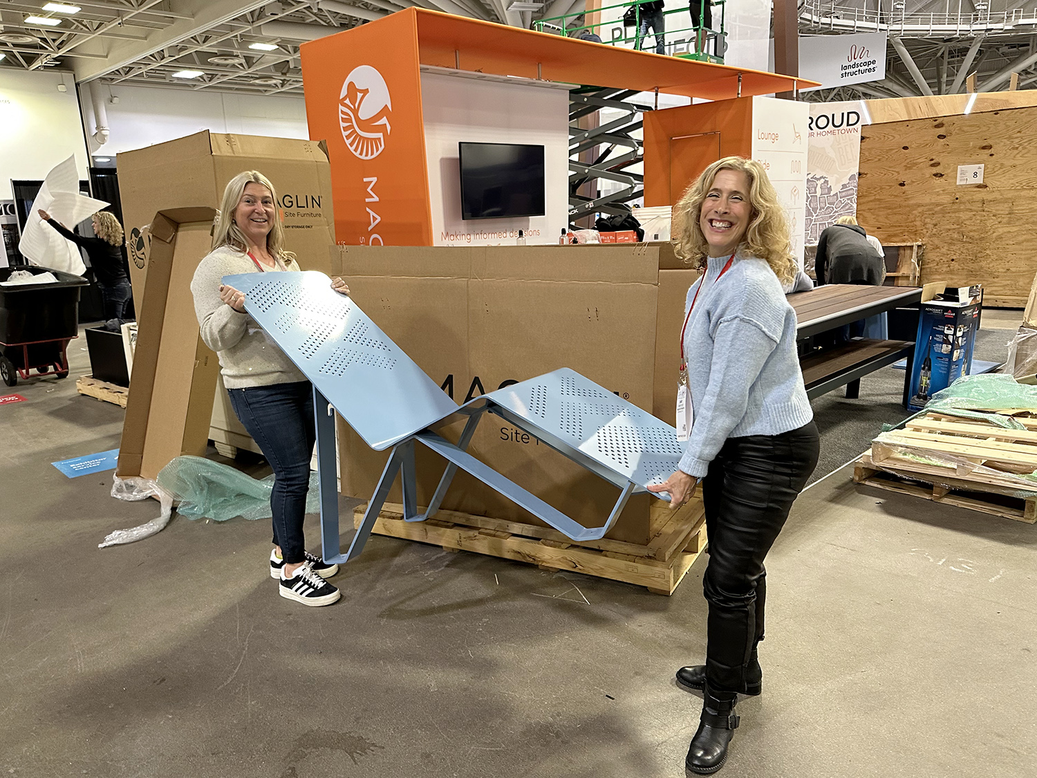 Two women are smiling and holding a metal lounge chair at an indoor event or trade show, surrounded by packaging materials and display booths.