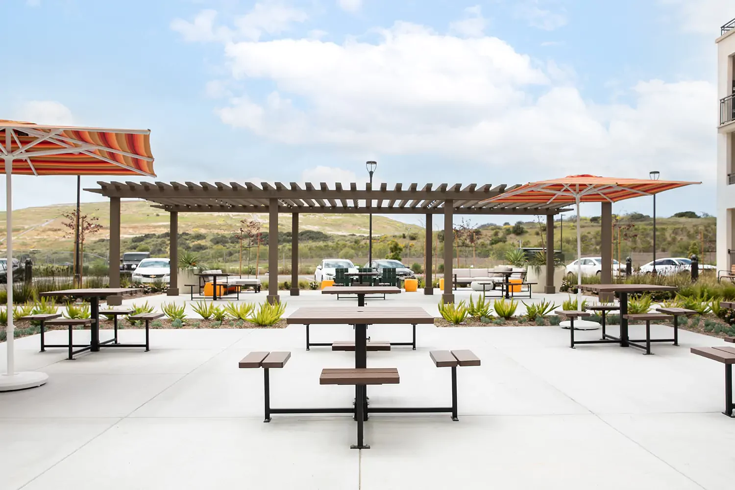 Outdoor patio area with wooden picnic tables, orange-striped umbrellas, a pergola, and landscaped plants, with parked cars and hills in the background under a partly cloudy sky.