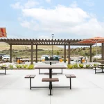 Outdoor patio area with wooden picnic tables, orange-striped umbrellas, a pergola, and landscaped plants, with parked cars and hills in the background under a partly cloudy sky.