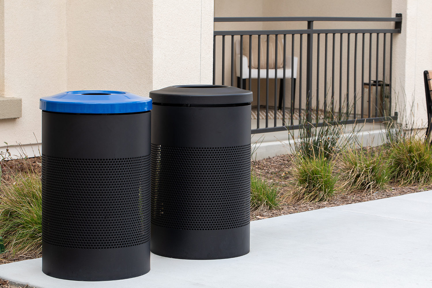 Two black outdoor trash bins, one with a blue lid and one with a black lid, stand on a concrete surface near a building and some landscaping.