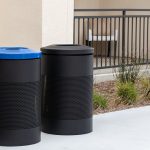 Two black outdoor trash bins, one with a blue lid and one with a black lid, stand on a concrete surface near a building and some landscaping.