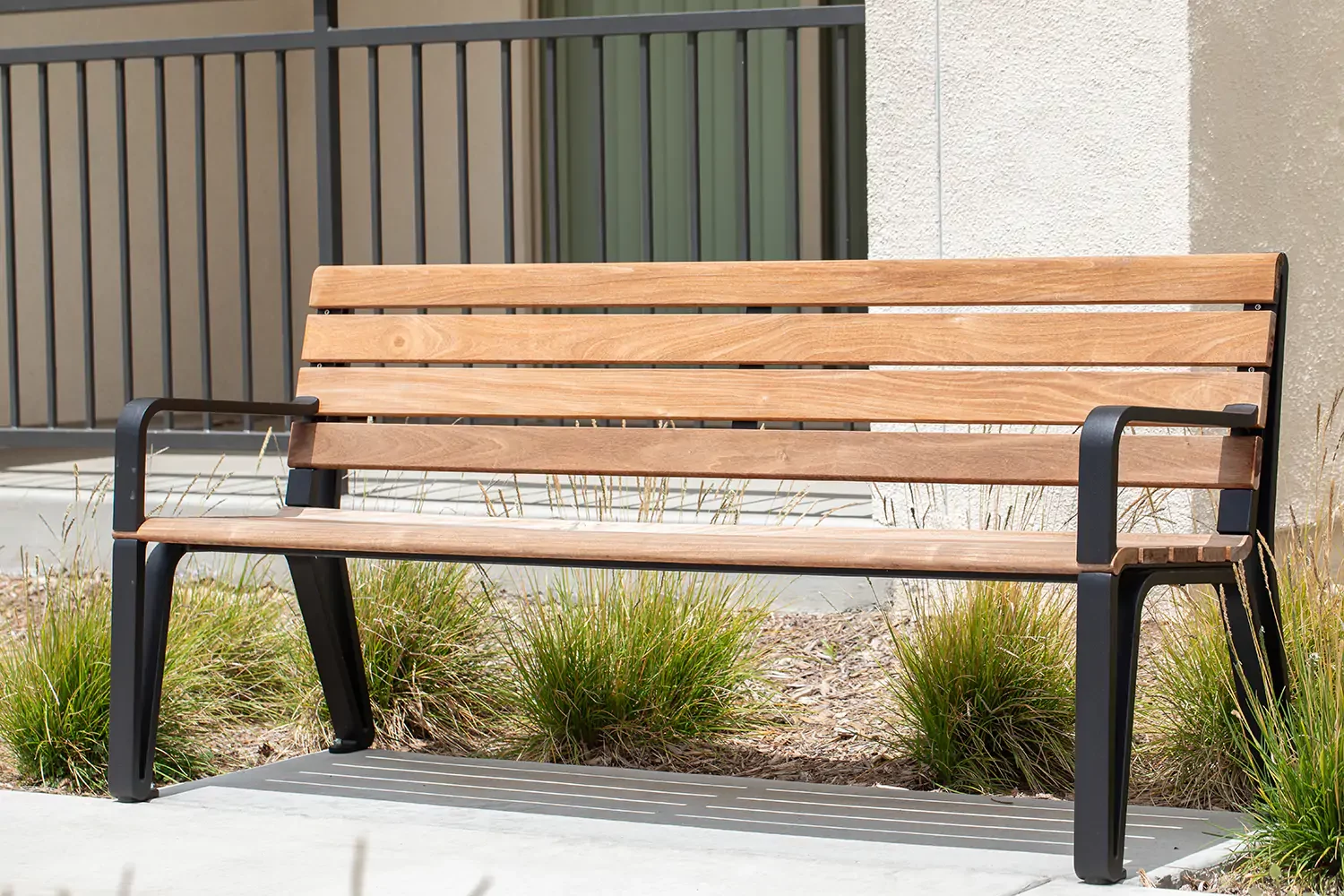 Un banc en bois avec des pieds et des accoudoirs en métal noir est placé sur une surface en béton près d'un bâtiment beige et de quelques herbes ornementales.