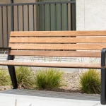 Un banc en bois avec des pieds et des accoudoirs en métal noir est placé sur une surface en béton près d'un bâtiment beige et de quelques herbes ornementales.