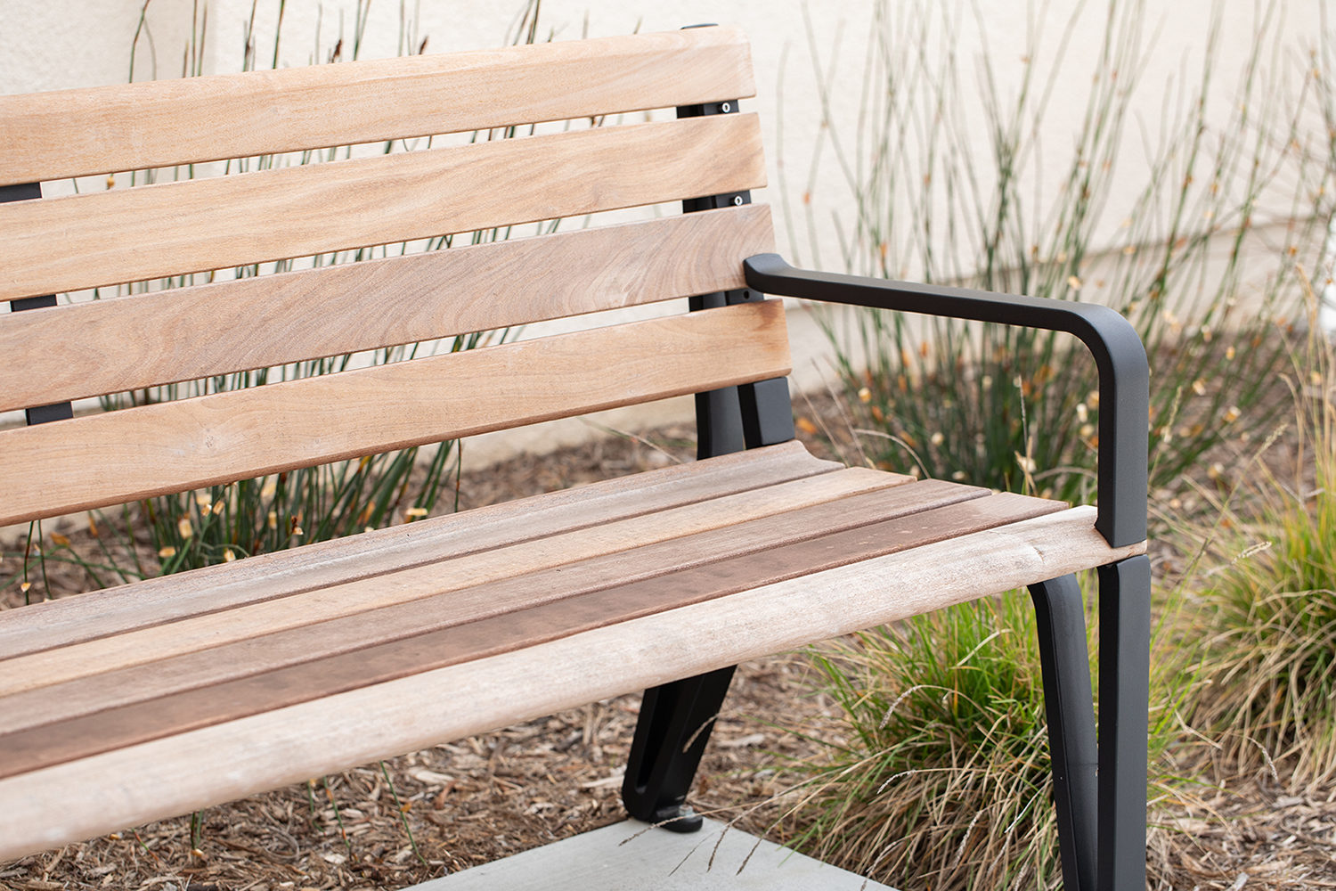 A partial view of the Maglin Iconic Collection bench with wood slats and black cast aluminum end arms sits in front of a stucco apartment building. Drought-tolerant grasses are planted behind the bench in a mulched bed.