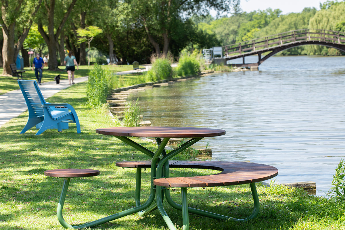 Maglin Fava Cluster Seating picnic table sits on the grass next to a river. There is a quaint bridge in the background as well as a pastel blue 720 Chair and people walking dogs on a path in the background as well.