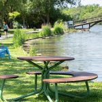 Maglin Fava Cluster Seating picnic table sits on the grass next to a river. There is a quaint bridge in the background as well as a pastel blue 720 Chair and people walking dogs on a path in the background as well.