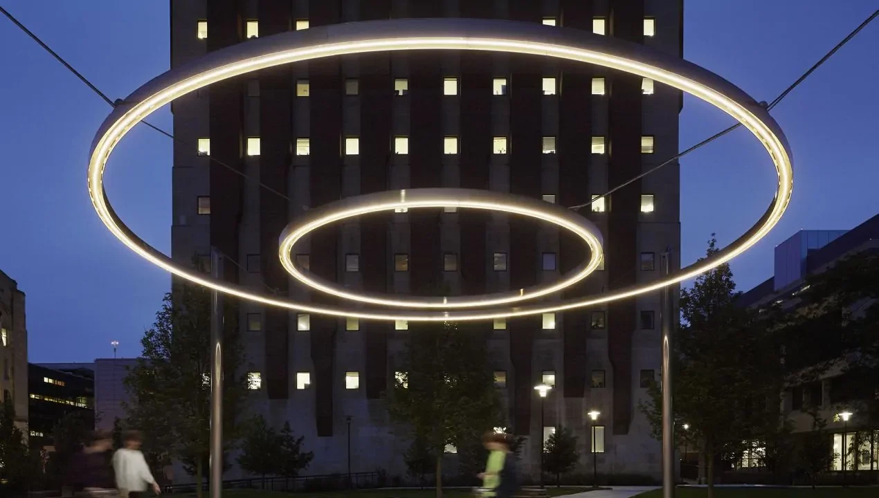 Two large, circular LED light installations are suspended outdoors at night, with a tall building and blurred pedestrians in the background.