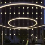 Two large, circular LED light installations are suspended outdoors at night, with a tall building and blurred pedestrians in the background.
