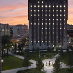A tall building with illuminated windows stands in a landscaped courtyard at sunset, with a few people gathered near a circular feature in the center.