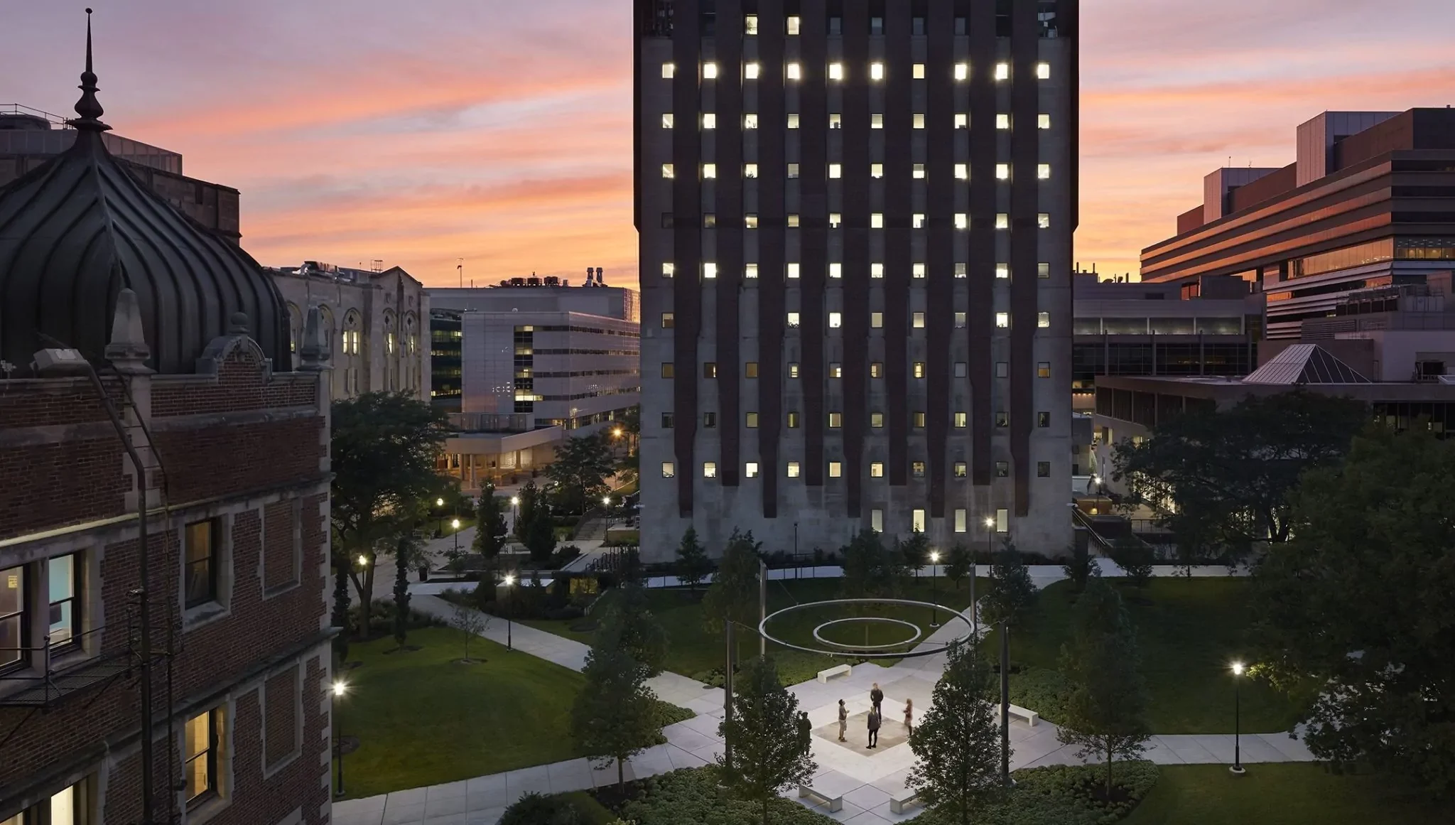 A tall building with illuminated windows stands in a landscaped courtyard at sunset, with a few people gathered near a circular feature in the center.