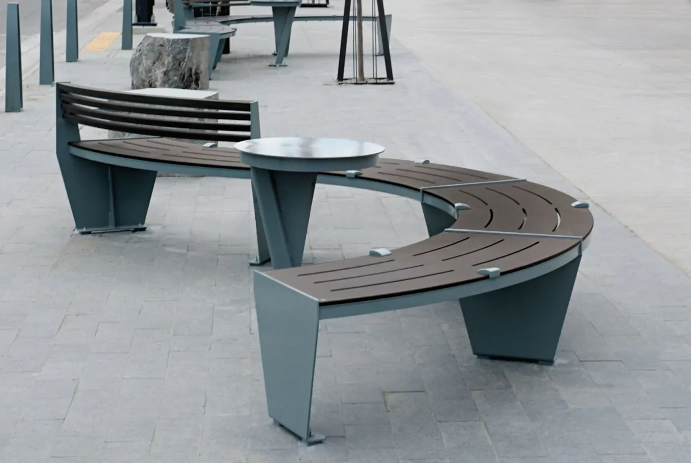Curved modern benches with metal legs and wooden slats surround a circular metal table on a paved outdoor area.