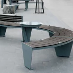 Curved modern benches with metal legs and wooden slats surround a circular metal table on a paved outdoor area.