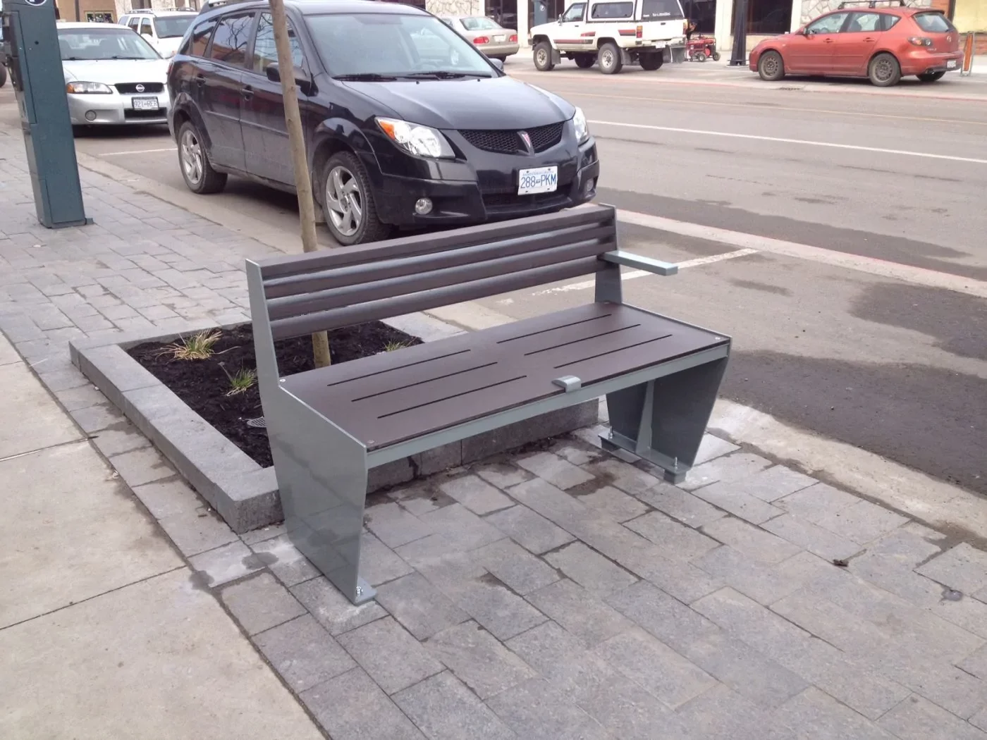 A metal and wooden bench with one armrest is placed on a paved sidewalk next to a small tree and parked cars on a city street.