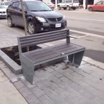 A metal and wooden bench with one armrest is placed on a paved sidewalk next to a small tree and parked cars on a city street.