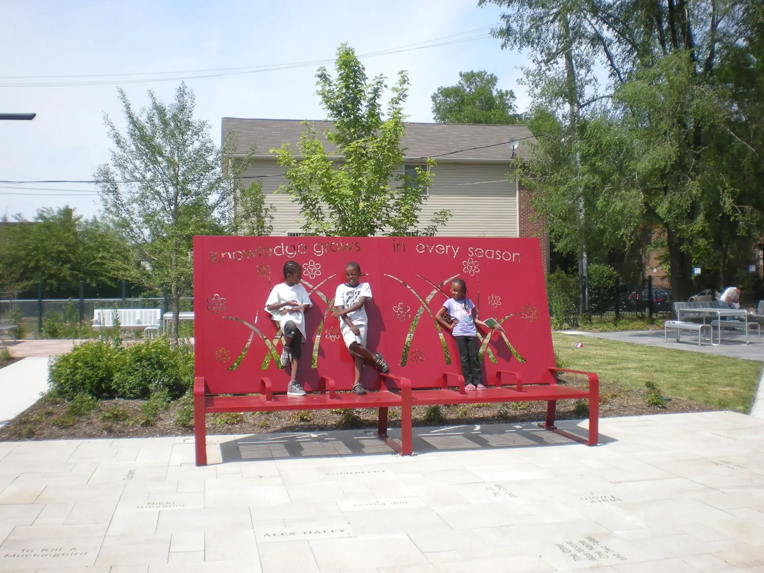 Three children stand on a red bench in front of a decorative red panel in a landscaped outdoor park area with trees and seating.