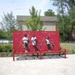 Three children stand on a red bench in front of a decorative red panel in a landscaped outdoor park area with trees and seating.