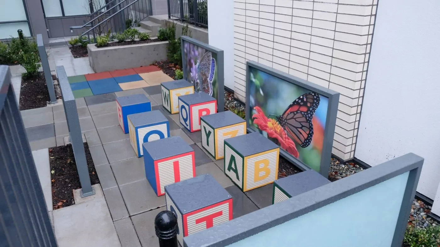 Large alphabet blocks are arranged in two rows on a paved outdoor area, with two framed butterfly photos displayed on the wall behind them.