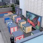 Large alphabet blocks are arranged in two rows on a paved outdoor area, with two framed butterfly photos displayed on the wall behind them.