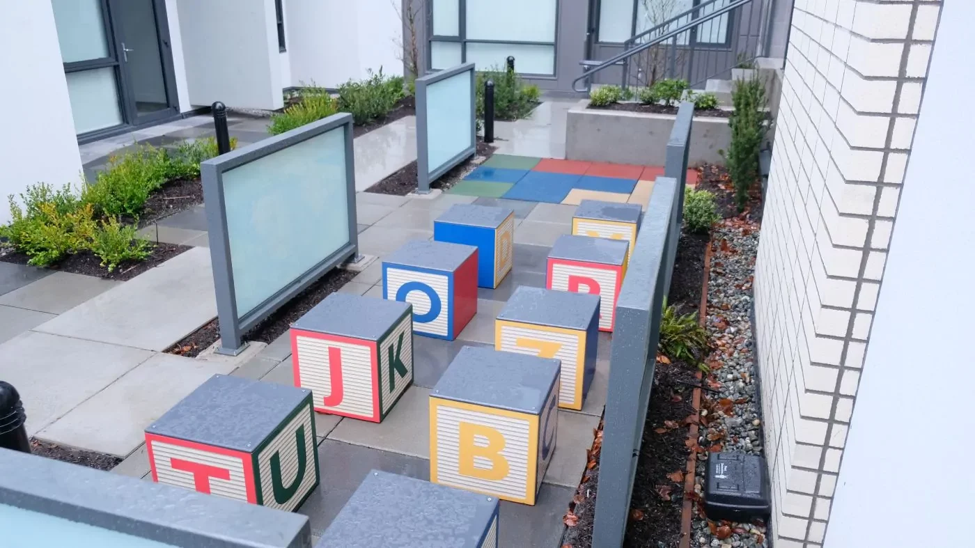 Colorful alphabet blocks are arranged in rows on a raised outdoor concrete platform next to a pathway with plants and a building in the background.