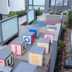 Colorful alphabet blocks are arranged in rows on a raised outdoor concrete platform next to a pathway with plants and a building in the background.
