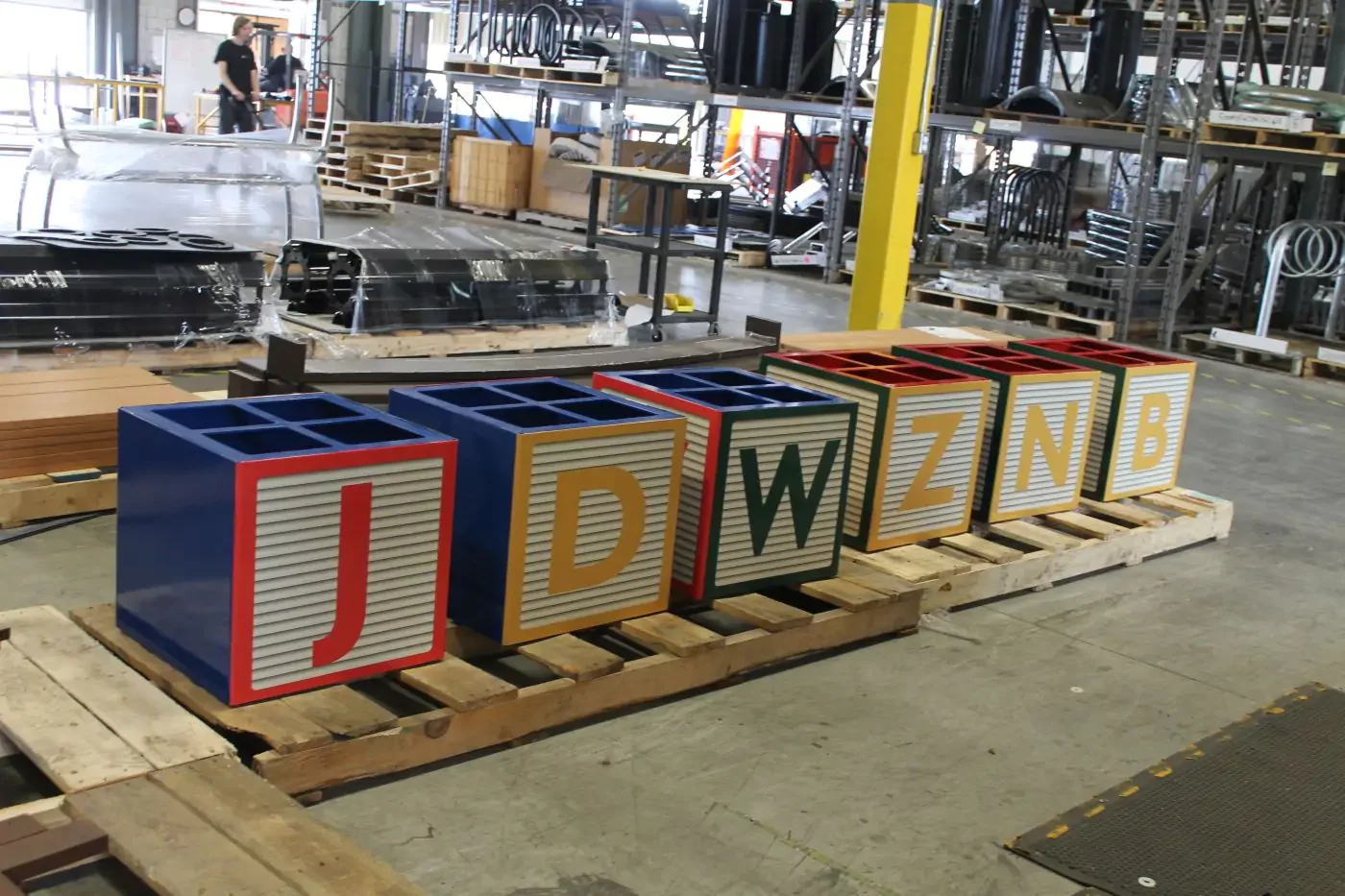 Six large, block-shaped containers with letters J, D, W, Z, N, and B sit on wooden pallets in an industrial warehouse setting.