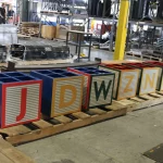 Six large, block-shaped containers with letters J, D, W, Z, N, and B sit on wooden pallets in an industrial warehouse setting.