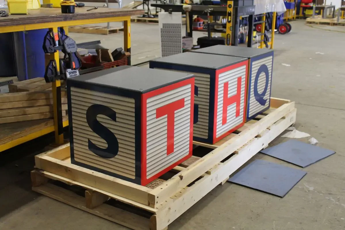 Three large cube blocks with the letters S, T, H, and Q on visible sides are placed on a wooden pallet in an industrial workshop setting.